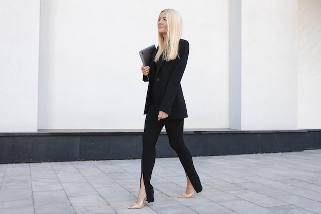 Une jeune femme en costume noir avec un ordinateur portable marche dans la rue