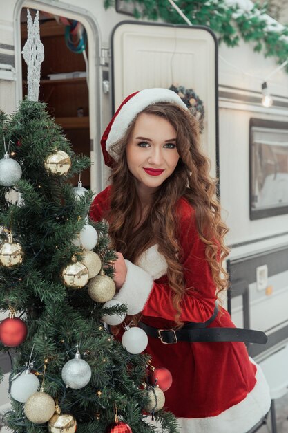 Une jeune femme en costume de père Noël décore le sapin de Noël au camping d'hiver en se préparant pour le