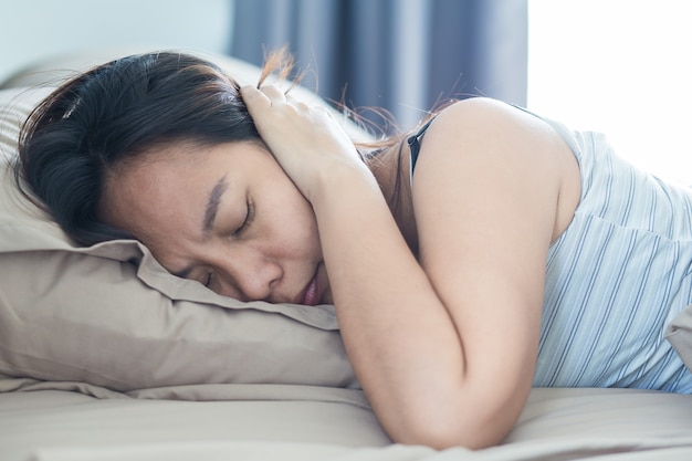 Jeune femme couchée dans son lit souffrant de son couvrant la tête et les oreilles avec oreiller