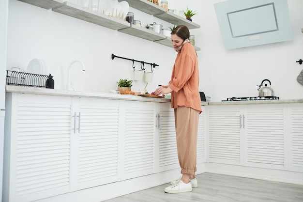 Jeune femme coupant des fruits au tableau et parlant au téléphone portable tout en se tenant dans la cuisine à la maison