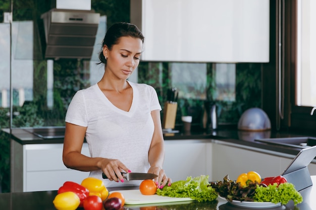 La jeune femme coupe des légumes dans la cuisine avec un couteau et un ordinateur portable sur la table