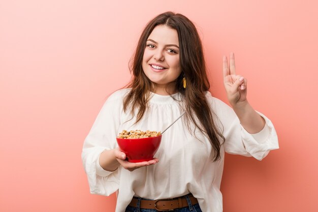 Jeune femme courbée taille plus tenant un bol de céréales montrant le signe de la victoire et souriant largement.