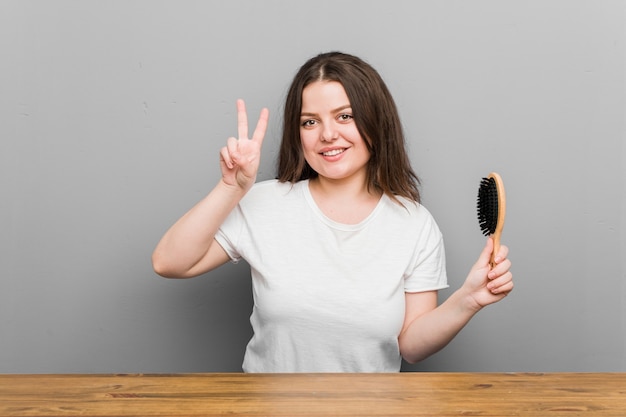 Photo jeune femme courbée de taille plus tenant une brosse à cheveux montrant le signe de la victoire et souriant largement.