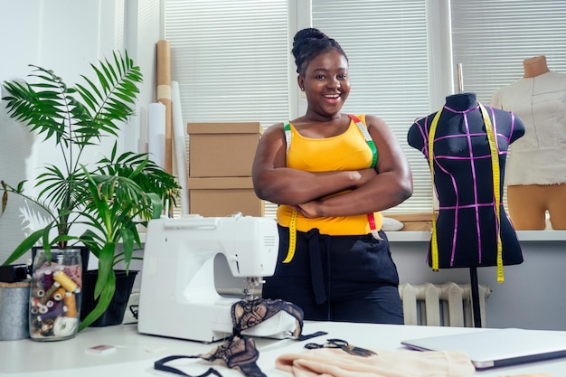 Jeune femme couturière afro-américaine coud des vêtements sur une machine à coudre au bureau de tailleur dans un style d'été tropical.