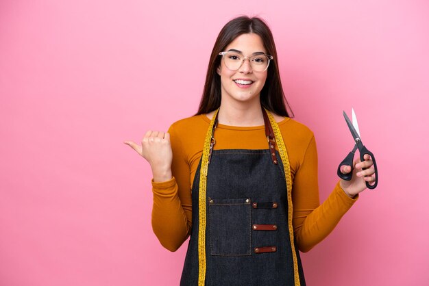 Jeune femme couturière brésilienne isolée sur fond rose pointant vers le côté pour présenter un produit