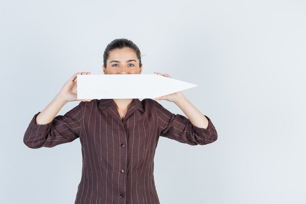 Jeune femme couvrant la bouche avec une affiche en papier en chemise, jeans et l'air curieux, vue de face.