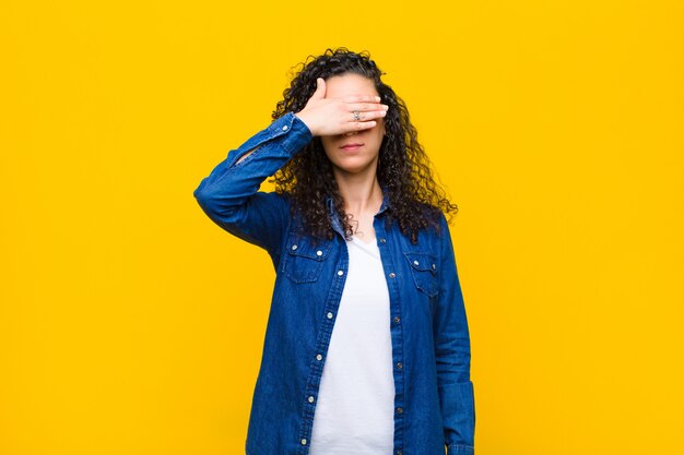 Jeune femme couvrant les yeux avec une main se sentant effrayé ou anxieux, se demandant ou attendant aveuglément une surprise sur le mur orange