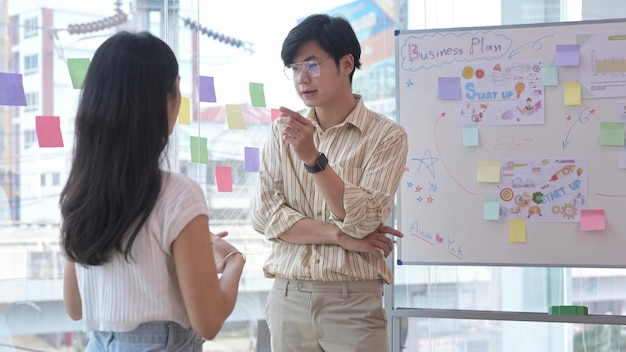 Jeune femme créative debout près d'un tableau à feuilles mobiles avec son collègue et discutant ensemble d'une nouvelle stratégie marketing