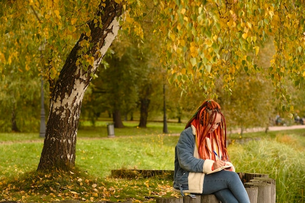 Jeune femme créative avec des dreadlocks dessin écrit dans un cahier assis sur des souches près du lac dans un parc