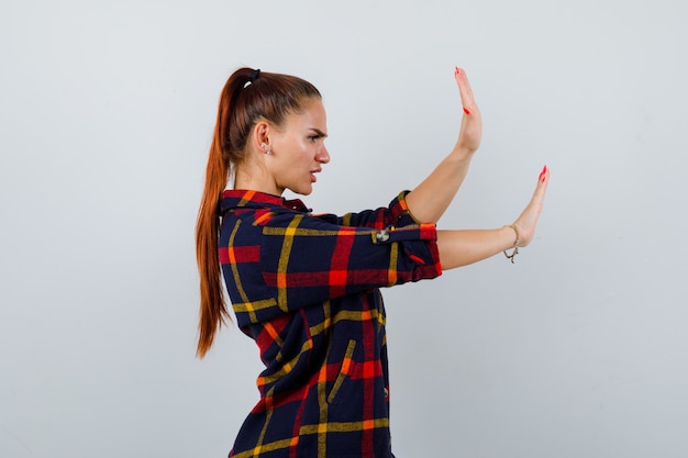 Photo jeune femme en crop top, chemise à carreaux, pantalon gardant les mains pour se défendre et ayant l'air effrayé, vue de face.