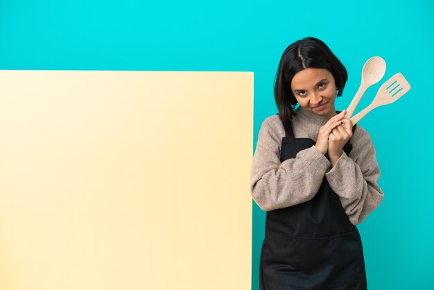 Jeune femme de cuisine métisse avec une grande pancarte isolée sur fond bleu maintient la paume ensemble. La personne demande quelque chose