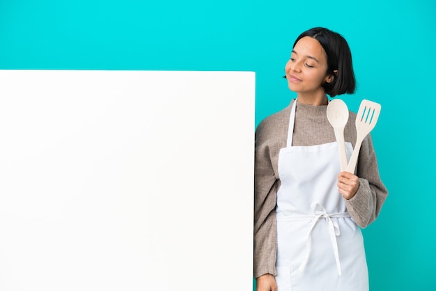 Jeune femme de cuisinier de race mixte avec une grande pancarte isolée sur fond bleu regardant sur le côté
