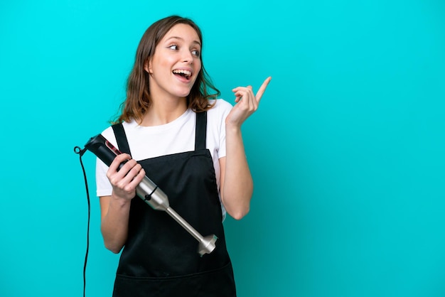 Jeune femme cuisinière caucasienne à l'aide d'un mélangeur à main isolé sur fond bleu dans l'intention de réaliser la solution tout en levant un doigt