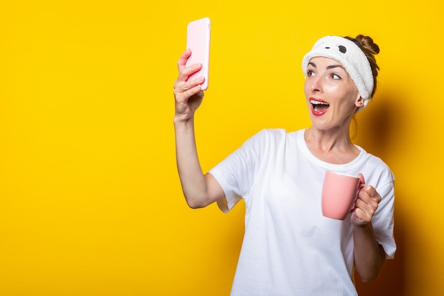 Jeune femme dans un bandage pour dormir et avec une tasse de café fait un selfie, posant sur un fond jaune.