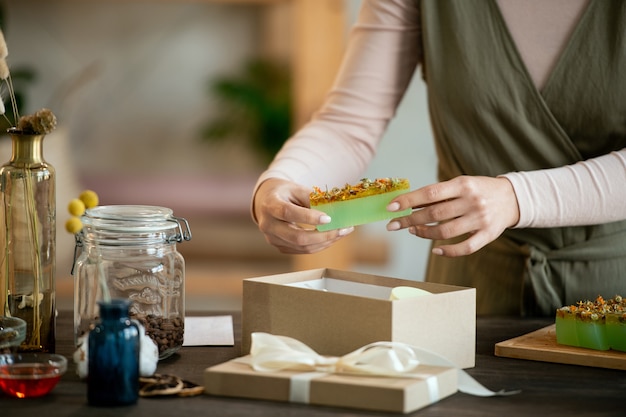 Jeune femme dans un bar d'emballage de vêtements décontractés de savon naturel frais fait à la main dans une boîte-cadeau en carton tout en préparant des cadeaux pour ses amis