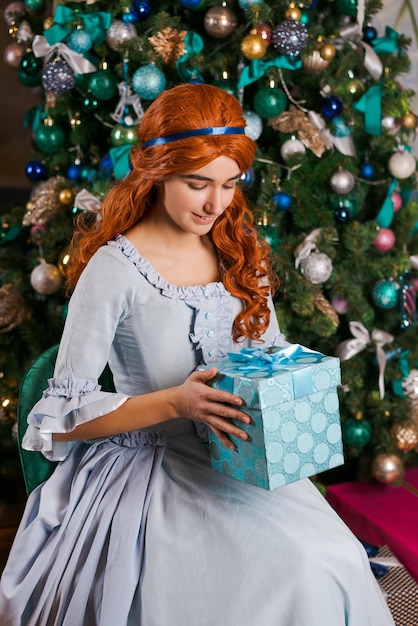 une jeune femme dans une belle robe est assise près d'un arbre de Noël tenant une boîte avec un cadeau dans ses mains