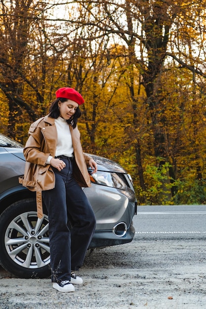 Une jeune femme dans un béret rouge se tient près de la voiture sur le fond d'une forêt d'automne