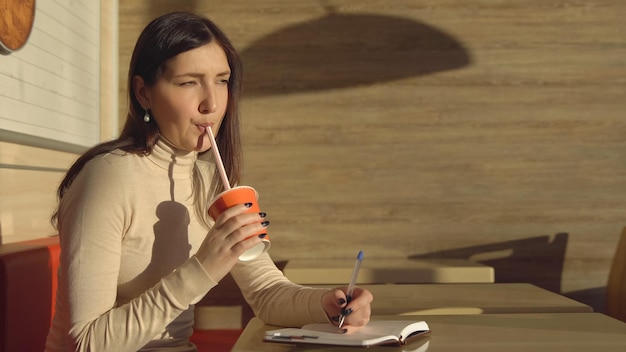 Une jeune femme dans un café prend des notes dans un cahier et boit du jus.