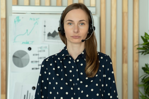 Jeune femme dans un casque debout dans un bureau travaillant comme centre d'appels ou opérateur de service d'assistance regarde la caméra