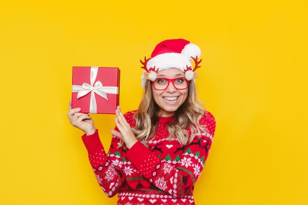 Une jeune femme dans un chandail de cerf rouge et un chapeau de Père Noël de Noël montre une boîte-cadeau sur fond jaune