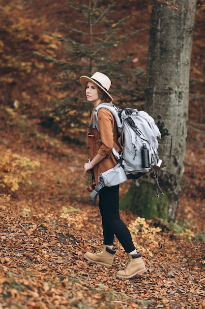 Jeune femme dans un chapeau élégant et un sac de voyage sur ses épaules, regardant autour de la charmante forêt d'automne