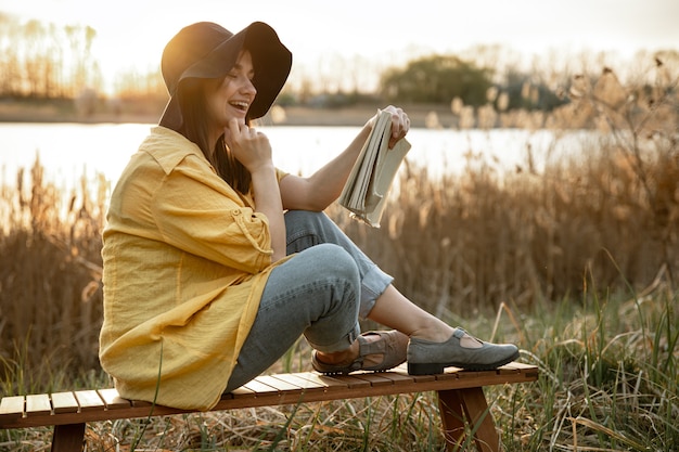 Photo une jeune femme dans un chapeau avec un sourire sur son visage lit un livre assis au bord de la rivière au coucher du soleil