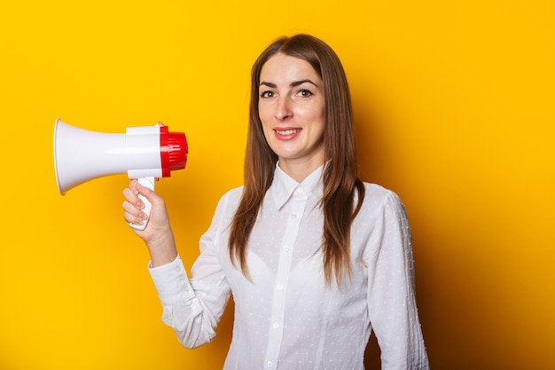 Jeune femme dans une chemise blanche tient un mégaphone dans ses mains sur un fond jaune