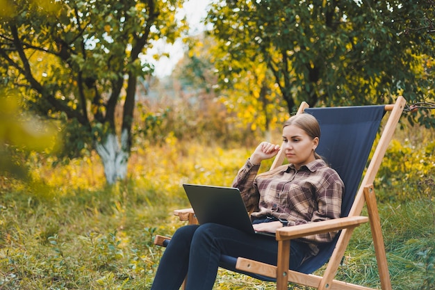 Jeune femme dans une chemise à carreaux assise sur une chaise à l'extérieur dans un jardin et travaillant sur un ordinateur portable