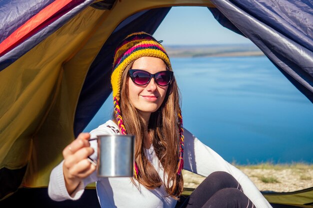 Jeune femme dans un drôle de chapeau du Népal assis dans un sac de couchage bleu dans la tente tenant une tasse thermos et buvant une tasse de café thé fond paysage lac. bonjour dans le voyage.