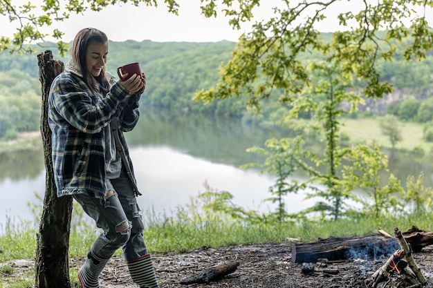 Une jeune femme dans la forêt près de la rivière se réchauffe au coin du feu et boit une boisson chaude