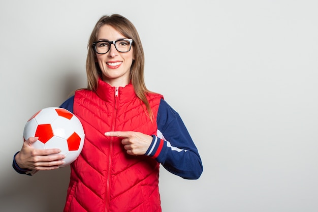 Photo jeune femme dans un gilet rouge tient un ballon de football dans ses mains et le montre avec son doigt