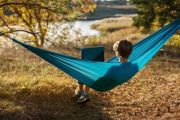 Jeune femme dans un hammok utilisant un ordinateur portable travaillant en plein air face au lac sur un concept d'opportunité d'emploi à distance au coucher du soleil