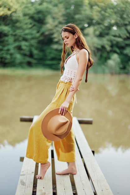 Une jeune femme dans un look hippie et ecodress dansant à l'extérieur au bord du lac portant un chapeau et un pantalon jaune au coucher du soleil d'été