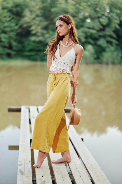 Une jeune femme dans un look hippie et ecodress dansant à l'extérieur au bord du lac portant un chapeau et un pantalon jaune au coucher du soleil d'été