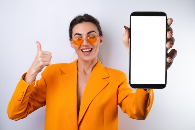 Une jeune femme dans des lunettes élégantes à la mode et une veste surdimensionnée orange vif sur fond blanc tient un téléphone avec un écran blanc vierge