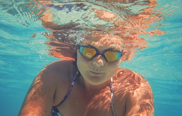 Jeune femme dans des lunettes de natation réfléchissantes nager sous l'eau en mer, des bulles éclairées par le soleil sur son visage, des détails sous-marins en gros plan