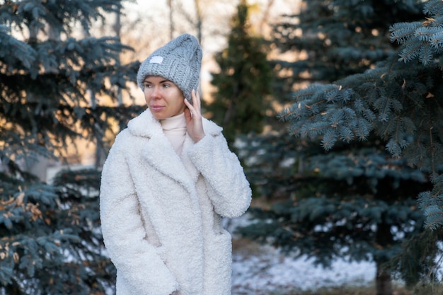 Une jeune femme dans un manteau de fourrure et un chapeau marche dans un parc d'hiver entre les arbres de Noël