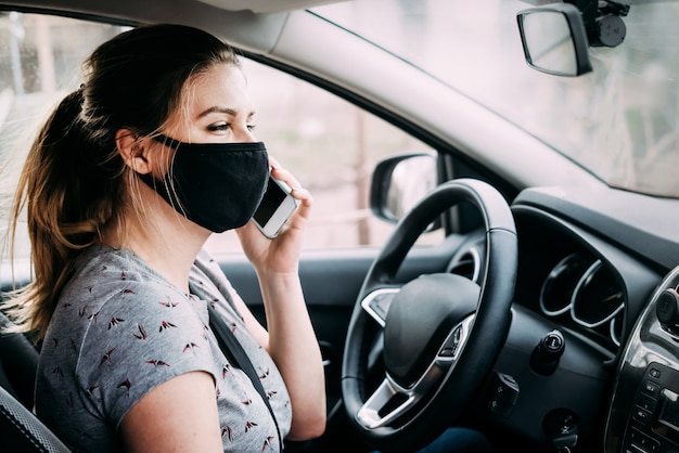 Jeune femme dans un masque médical assis dans une voiture avec conduite à gauche et parler au téléphone