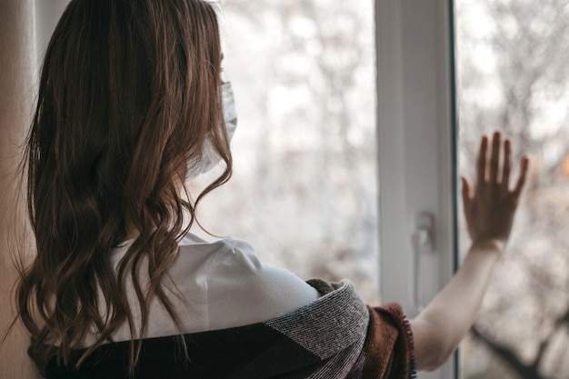 Jeune femme dans un masque médical regarde par la fenêtre et tient sa main sur la vitre, la quarantaine, l'isolement, le coronavirus, rester en sécurité à la maison,