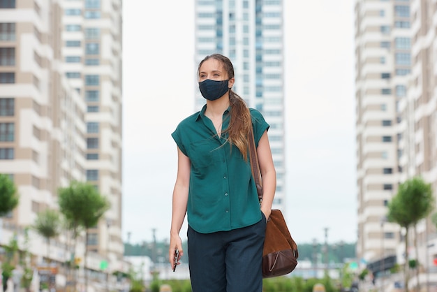 Une jeune femme dans un masque médical tenant un smartphone et enfonçant une autre main dans une poche de pantalon marche