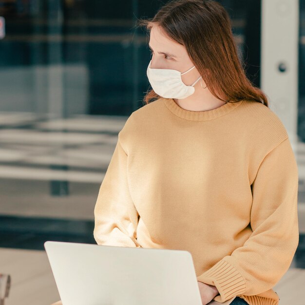 Jeune femme dans un masque de protection à l'aide d'un ordinateur portable dans une rue de la ville