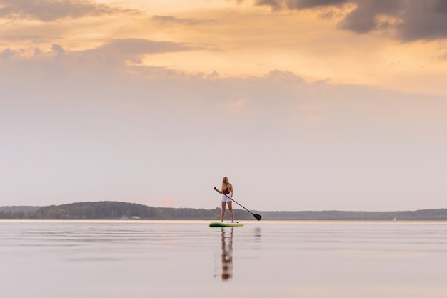 Photo jeune femme dans la mer se lever sur la planche de paddle sup avec de belles couleurs de coucher ou lever de soleil