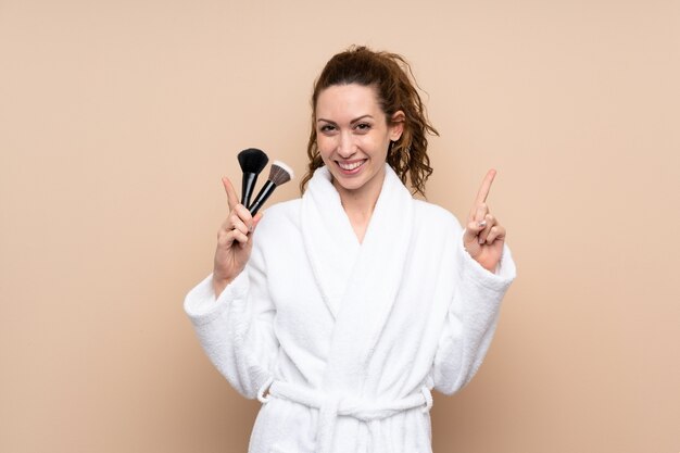 Jeune femme dans un peignoir tenant des pinceaux de maquillage pointant vers le haut une bonne idée