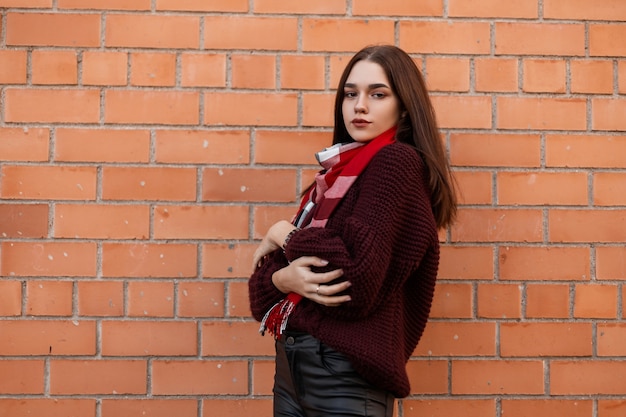 Jeune femme dans un pull bordeaux et un pantalon en cuir noir se dresse dans la ville près d'un mur de briques