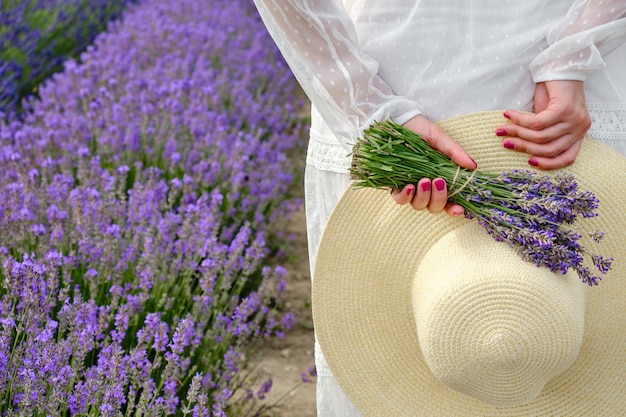 Jeune femme dans une robe de lumière blanche tenant derrière son dos un chapeau et un bouquet de lavande