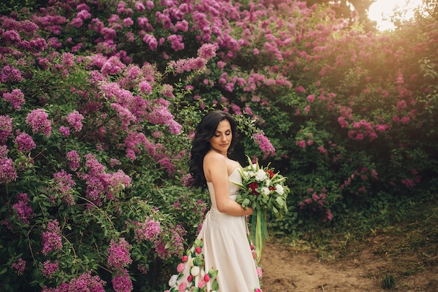 Jeune femme dans une robe de pivoine se dresse dans un jardin lilas fleuri au coucher du soleil