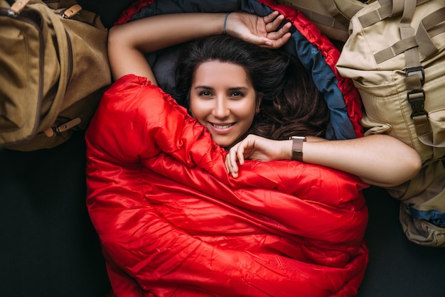 Une jeune femme dans un sac de couchage confortable dans une tente, vue de dessus. Un touriste dans un sac de couchage. Un voyageur enveloppé dans un sac de couchage rouge. Voyage, concept de camping, aventure. Voyager avec une tente