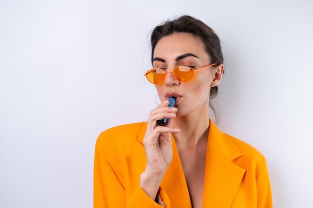 Une Jeune Femme Dans Des Verres élégants à La Mode Et Une Veste Surdimensionnée Orange Vif Sur Fond Blanc Fume Une Cigarette électronique