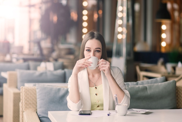 Jeune femme dans une veste blanche travaillant dans un café avec des papiers, buvant du café