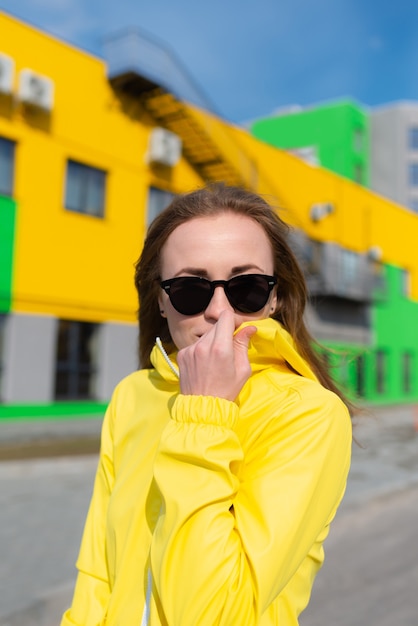 Une jeune femme dans une veste jaune et des sungalsses avec des bâtiments de couleurs vives en arrière-plan